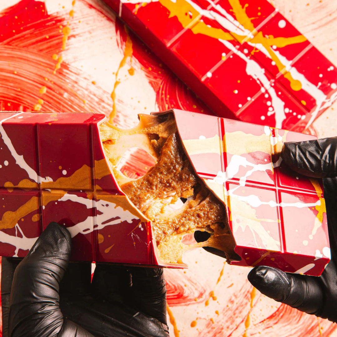 A person holding a broken piece of chocolate slab, with the slab itself having a red and yellow marbled design, indicating a festive appearance.