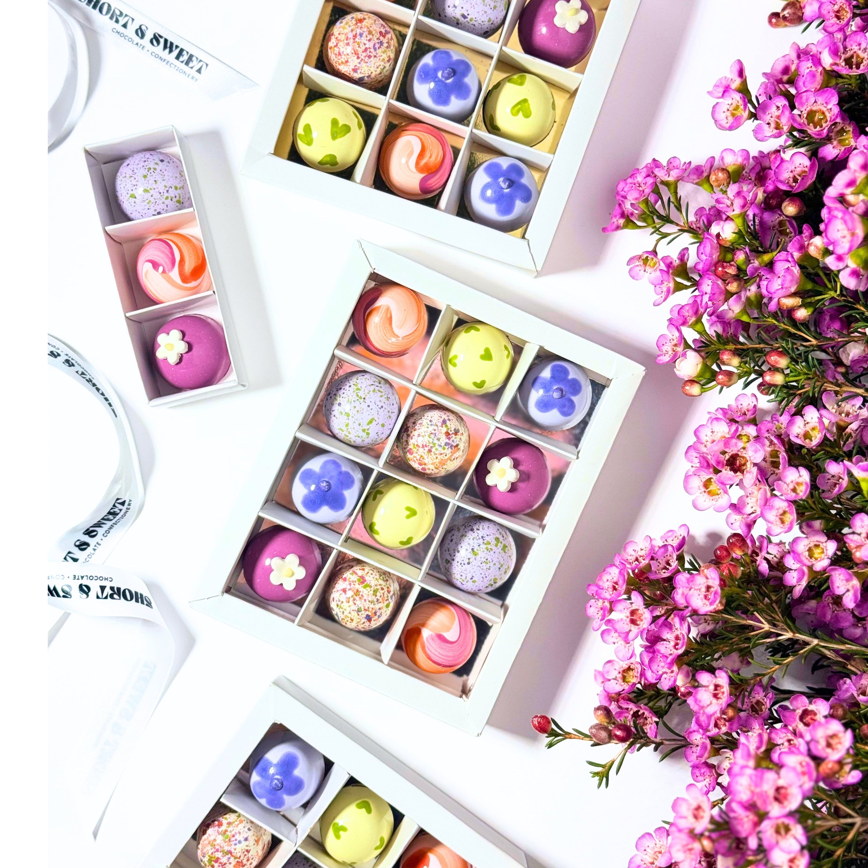 White boxes of assorted colorful bonbons arranged on a white surface, styled with pink flowers and a white ribbon.