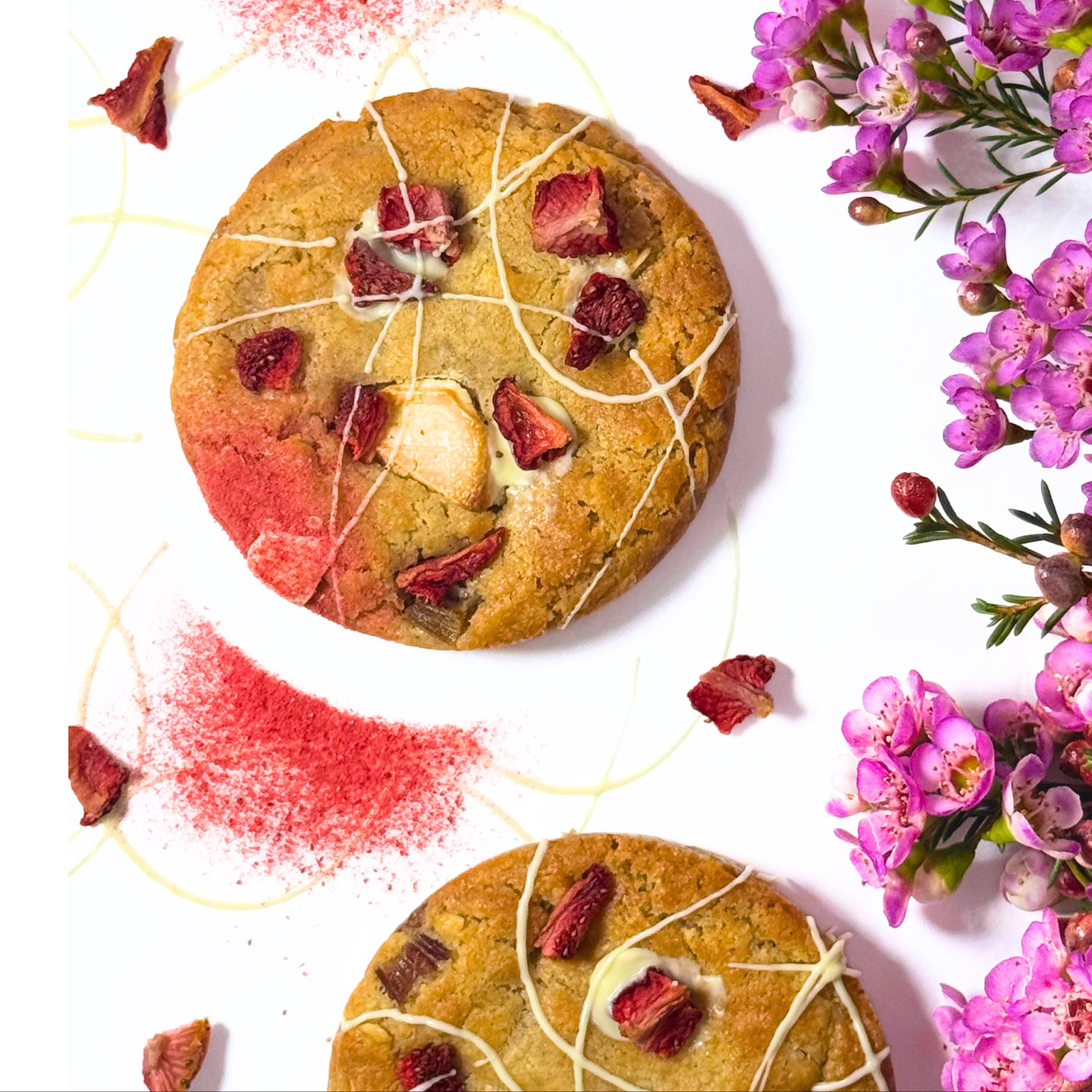 Two round chocolate chip cookies topped with dried strawberries and white chocolate drizzle, arranged on a white background with pink flowers and red powder accents.