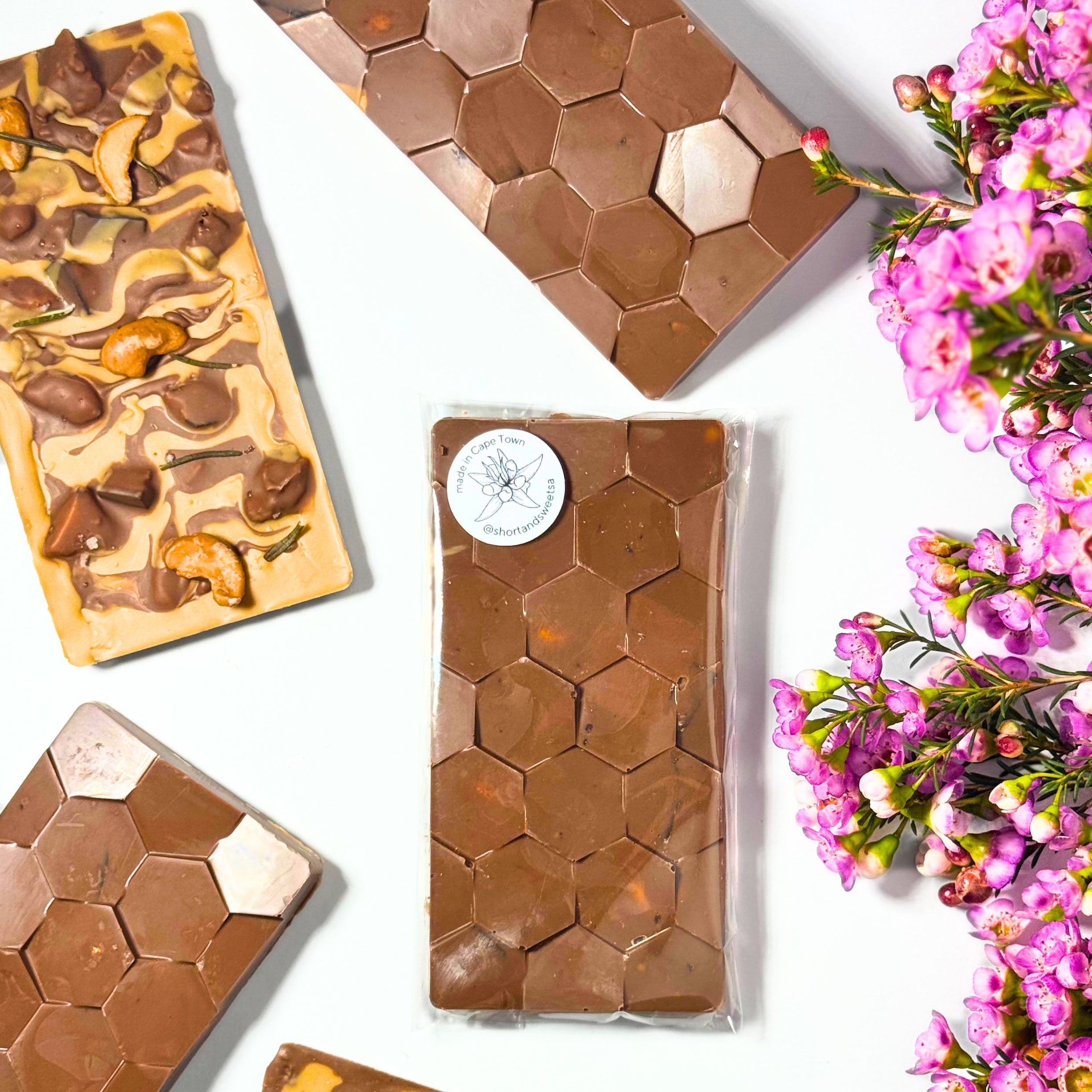 Rectangular bars of chocolate with swirls of dark and milk chocolate, topped with honeycomb and cahew nut pieces, photographed on a white background with pink flowers along the right side.