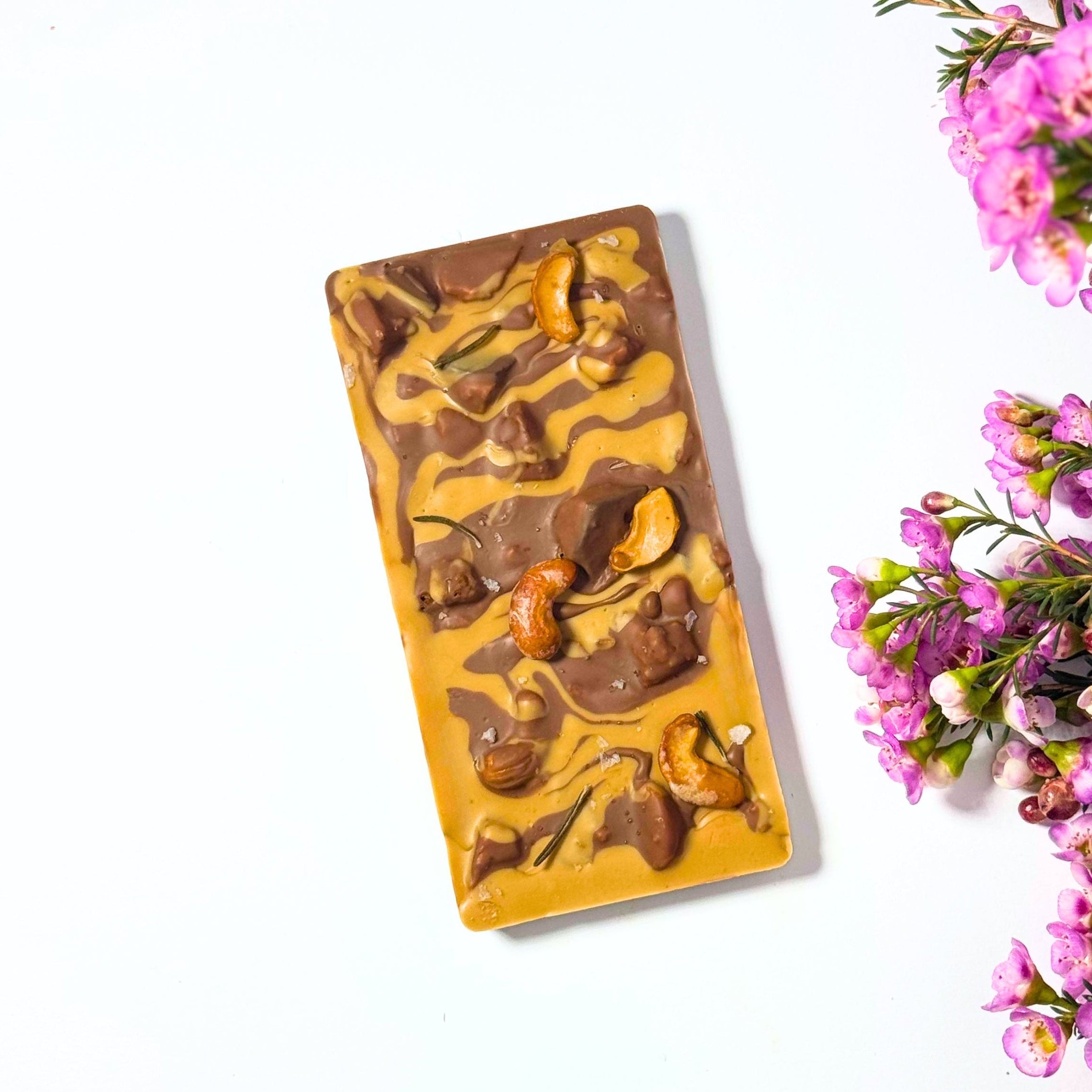 A rectangular bar of chocolate with swirls of dark and milk chocolate, topped with honeycomb and cahew nut pieces, photographed on a white background with pink flowers along the right side.