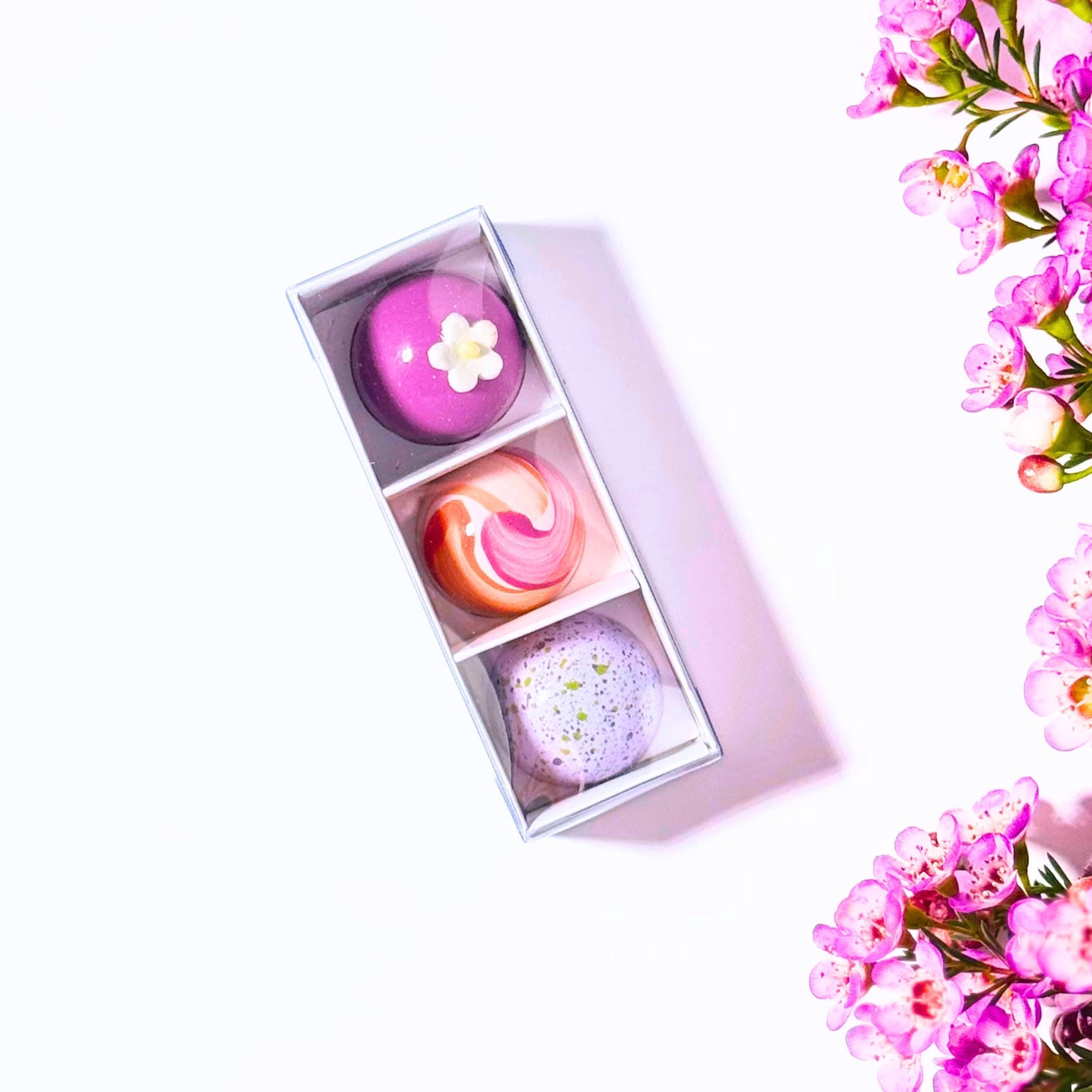A small rectangular box containing three colourful bonbons placed on a clean white background with pink flowers arranged along the right side of the frame.