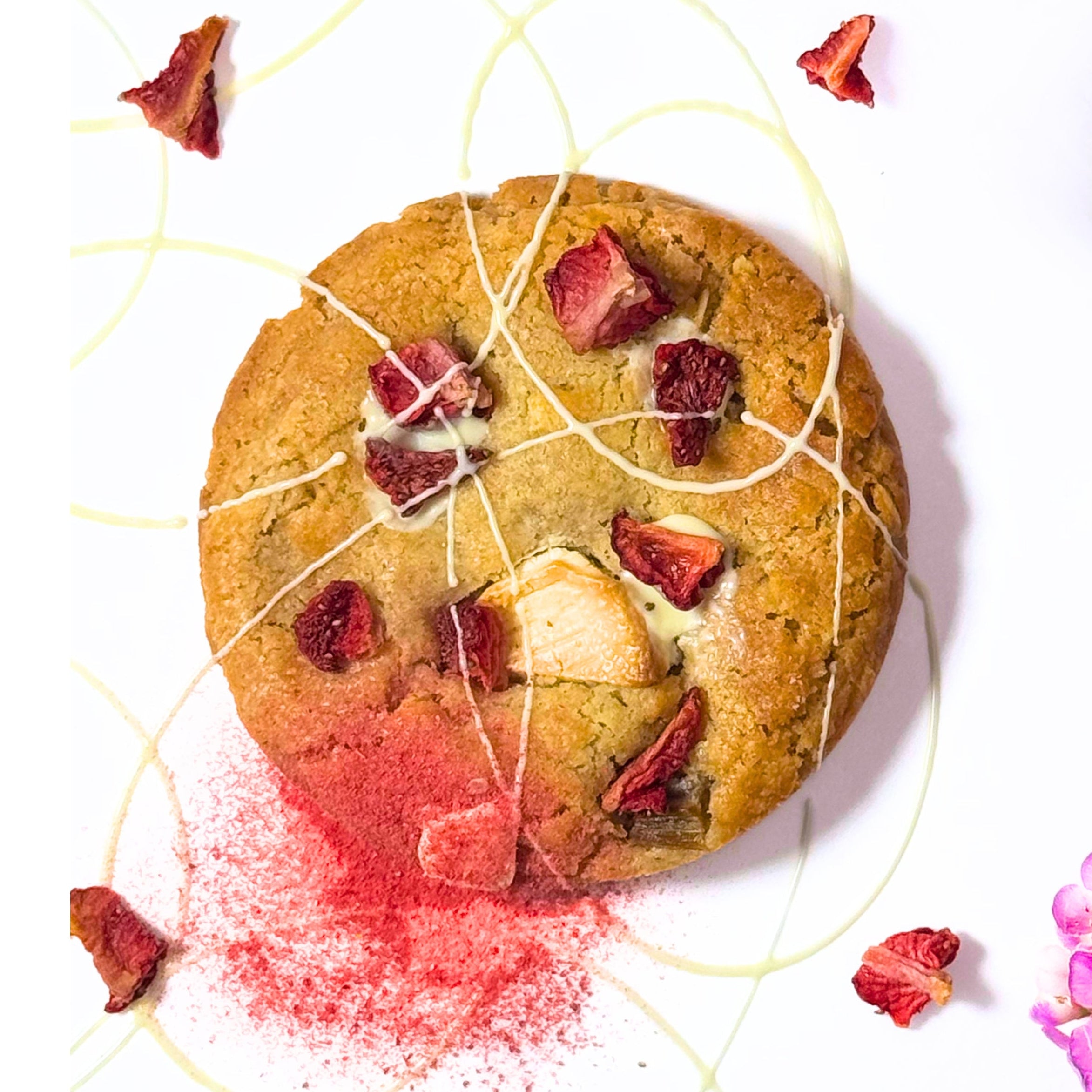 A round chocolate chip cookie topped with dried strawberries and white drizzle, styled on a white background with pink flowers and red powder scattered near it.