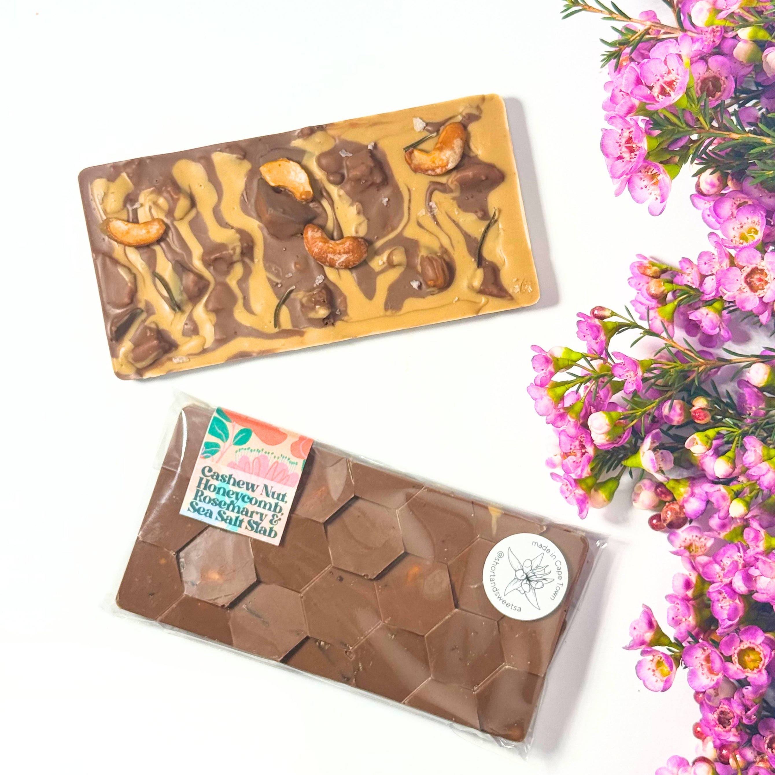 Two artisan chocolate bars displayed on a white background next to pink flowers, with swirls and cashew pieces.