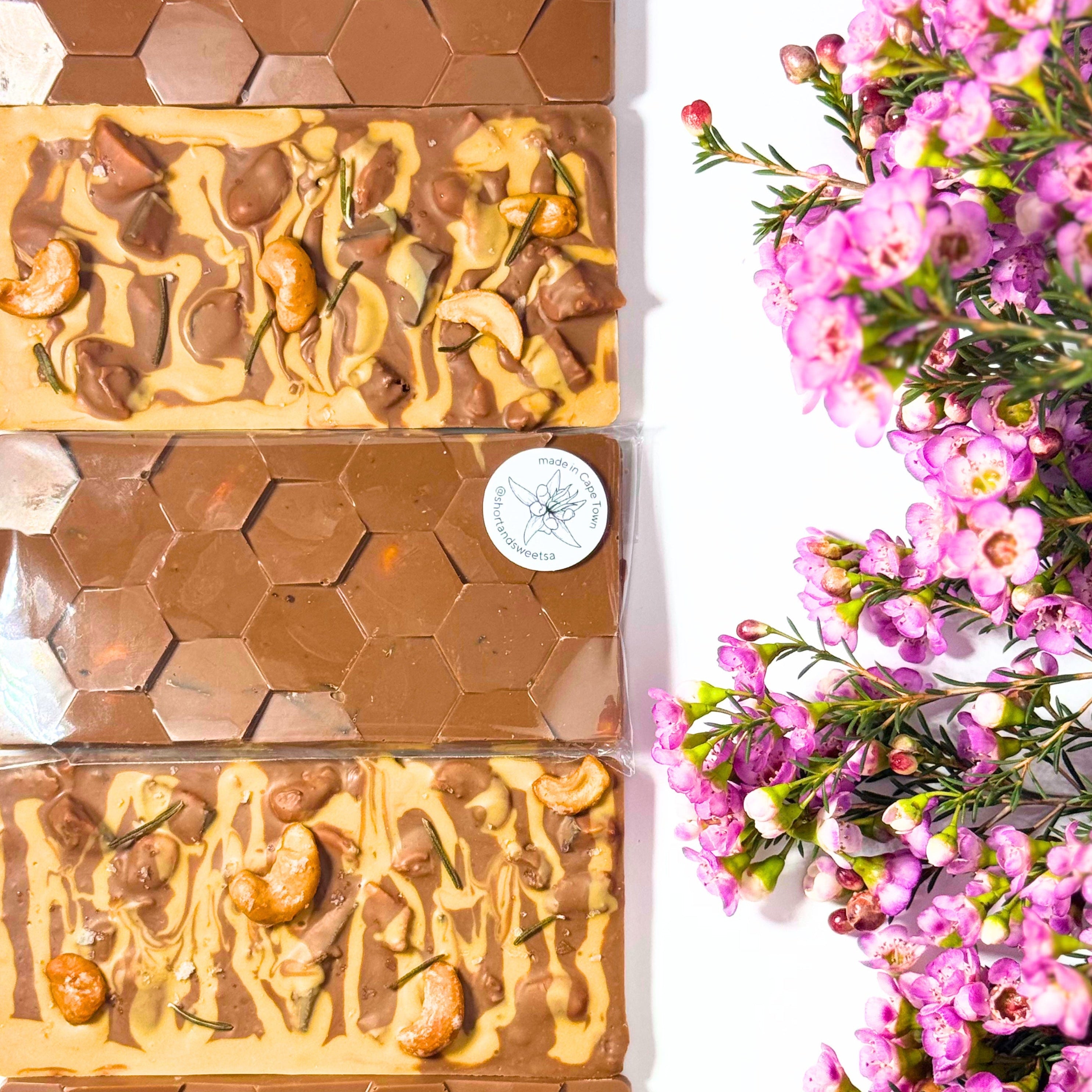 Four rectangular bars of chocolate arranged vertically on a white background, alternating between smooth dark brown hexagonal chocolate and milk chocolate with cashews, honeycomb, and drizzles. Pink flowers are placed along the right side of the frame.